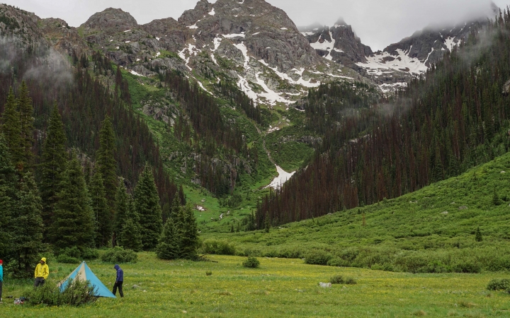 A tent sits in a green valley surrounded by mountains. Three people are standing near the tent.